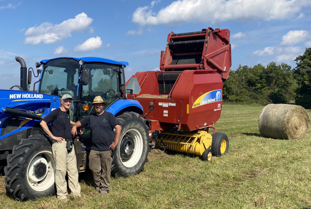 Birder and farmer stand in hayfield with farm equipment and hay bale